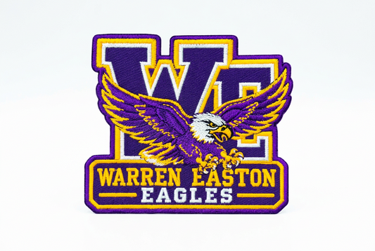 An embroidered Warren Easton Eagles patch featuring a purple and gold eagle with a white head, set against a purple and gold 'WE' monogram background.